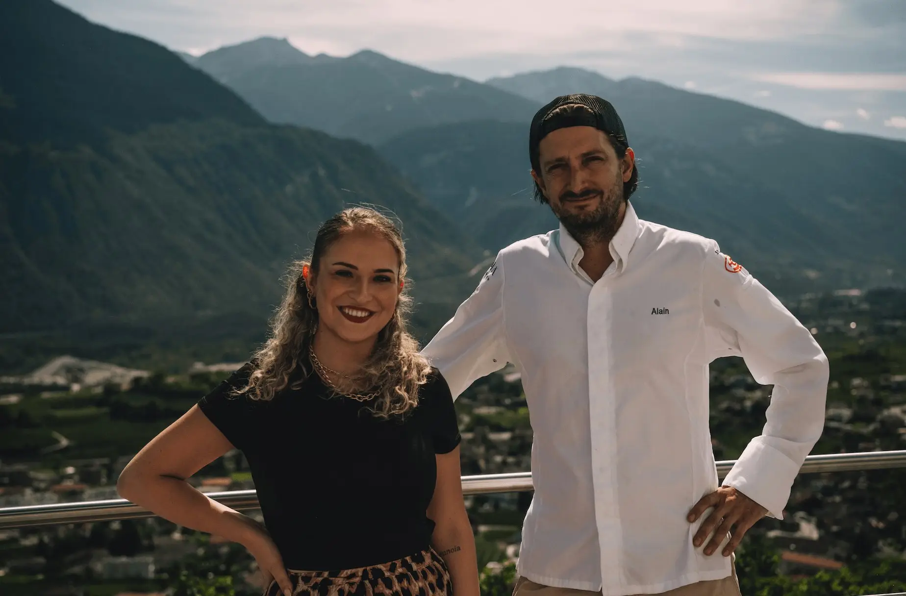 Alisha Cina et Alain Lerjen en veste de cuisine, derrière eux les montagnes au-dessus de la vallée du Rhône